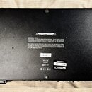 Fotografie zachycuje spodní stranu černého elektronického zařízení. Na spodní straně jsou dvě nálepky s texty: jedna obsahuje varovné doporučení „WAR…