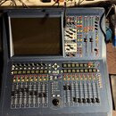 Modrý digitální mixovací pult s velkým displejem v levé části. Na ovládacím panelu jsou vidět posuvníky, tlačítka a vpravo sekce s faderem. Uprostřed…