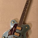 Kytara s polodupatým tělem v modrozelené barvě. Na hlavě a na těle je vidět nápis GRETSCH. Nástroj má dva snímače, červený pickguard a stříbrný hnízd…