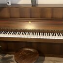 Hnědé dřevěné piano s bílými a černými klávesami. Nástroj je doplněn kulatou dřevěnou stoličkou v podobném odstínu. Na povrchu nejsou viditelné žádné…