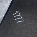 Detailní záběr na černý, texturovaný povrch s ručně napsaným bílým číslem 1777. Předmět je zasazen do světle šedého, kovového rámu s viditelnou koroz…