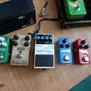 Pět kytarových efektů v řadě na dřevěném povrchu. Zleva jsou to zelený pedál CORONA od tc electronic, zlatý MXR CUSTOM BADASS Modified O.D., bílý BOS…