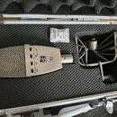 V otevřeném hliníkovém kufru s černou pěnovou výplní je umístěn stříbrný mikrofon a kovový shock mount. Na těle mikrofonu je vidět logo SE a označení…