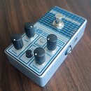 Kytarový efekt v hliníkovém šasi s modrým vzorovaným horním panelem. Na panelu jsou čtyři černé otočné knoflíky s popisky bass, gain, treble a volum.…