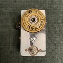Kytarový efekt pedál s kovovým stříbrným šasi. Na horní části je umístěno velké žluté kolečko s nápisem TONE WHEEL GUITAR PEDAL SERIES BY SALVATION A…