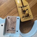 Fotografie zachicuje hřifoň a tělo elektrické kytary v modré barvě. Na dřevěné hlavě hřifu je vyryto logo s postavou kytaristy a nápisy "WARMOTH", "L…