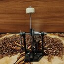 Černý bicí pedál s kovovými součástmi a bílým tlumicím polštářkem na horní části. Na základní desce je viditelný nápis "TAMA". Povrch nástroje vykazu…