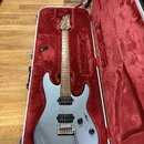 Stříbrná elektrická kytara v otevřeném tvrdém kufru s červeným vnitřkem. Na hlavě hředu je bílý nápis Ibanez. Kytara má dva humbuckery, dva ovládací…