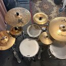 Kompletní bicí souprava s několika činely a bubny. Na činelech jsou viditelné nápisy Zildjian, CUSTOM a jedno velké písmeno K. Bubny mají napnuté mem…