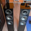 Dva vysoké stojací reproduktory v černém lesklém provedení s třemi měničy každý. V pozadí na bílé židli stojí jeden menší regálový reproduktor v podo…
