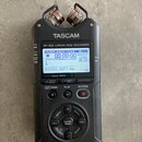 Černý digitální nahrávač s nápisy TASCAM a DR-40X Linear PCM RECORDER. Zařízení má LCD displej s zobrazením času a vlnové formy, horní mikrofony a ov…