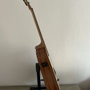 Akustická kytara s přírodním dřevěným povrchem, umístěná na černém stojanu. Na boční straně korpusu je vidět černý vstup pro kabel. Nástroj nemá žádn…