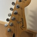 Detail hlavy elektrické kytary z přírodního dřeva s chromovanými ladičkami. Na dřevě jsou čitelné nápisy Fender®, STRATOCASTER® a ORIGINAL Contour Bo…