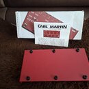 Fotografie zobrazuje červený kovový pedalboard s gumovými nožičkami. Vedle něj je položen manuál s nápisem CARL MARTIN MANUAL OCTA-SWITCH MK3 a krabi…