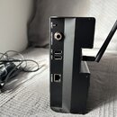 Černý elektronický přístroj s připojenou anténou a napájecím adaptérem. Na zadní straně jsou viditelné nápisy RESET, FOOTSWITCH, USB 1, USB 2 a ETHER…