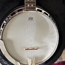 Bílé banjo v černém kufru s chromovaným kováním. Na bláně je vidět logo REMO a nápis WEATHERKING BANJO HEAD ONLY. Hmatník má ozdobné vložky a na spod…
