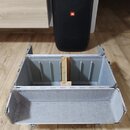 Otevřený hliníkový přepravní kufr s šedým vnitřním polstrováním. Uvnitř se nachází jedna dřevěná příčka. V pozadí je vidět černý reproduktor s červen…