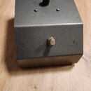 Šedý kovový box s textem Voice box na horní straně. Na vrchu je umístěn jeden hnědý konektor a čtyři malé šrouby. Na přední straně se nachází jeden k…