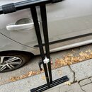 Černý kovový stojan na klavír nebo syntezizér s horní lištou a dvěma spodními oporami. Na spodní části je viditelné malé zlaté logo, které není čitel…