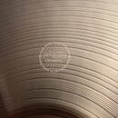 Detailní záběr na povrch kovového činely s koncentrickými rýhami. Uprostřed je vyryto logo Paiste a text HAND MADE IN SWITZERLAND v kruhovém rozložen…