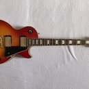 Elektrická kytara s tělem v červeně-žlutém sunburst provedení a černým pickguardem. Na hlavě je bílé logo Epiphone a nápis Les Paul Custom. Hриф má v…