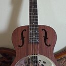 Hnědá akustická kytara typu resonator s kovovým, chromovaným krytem v těle. Na hlavě hředu je bílý nápis Harley Benton a na krku visí oranžová visačk…