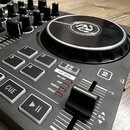 Černý DJ controller s jog-colem, které má v centru bílé logo. Na ovládacím panelu jsou viditelné tlačítka s nápisy CUE, PAD MODE, HOT CUE, LOOP, SAMP…