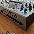 Stříbrný elektronický sekvenční přístroj s klávesami a ovládacími knoflíky. Na přední straně je velký nápis Roland MC-505 a v dolní části jsou vidite…