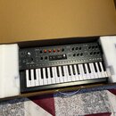 Černý syntetizátor s klaviatura v kartónovém boxu s polystyrenovými výplněmi. Na ovládacím panelu jsou viditelné nápisy OSCILLATORS, LFO, CYCLING ENV…