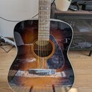 Akustická kytara s tělem v provedení sunburst (přechod od žluté k tmavě hnědé/černé). Nástroj má tmavý hrací hradík atmavě červený vzorovaný pickguar…
