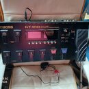 Černý kytarový procesor s nápisy BOSS a GT-100 EFFECTS PROCESSOR. Zařízení obsahuje displej, několik ovládacích tlačítek a pět stupňovitých spínáčů o…