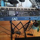 Klaviatura v tmavě modré barvě umístěná na černém X-stojanu. Na přední straně je bílý nápis POLYBRUTE, logo (A) v kruhu a značka ARTURIA. Přístroj má…