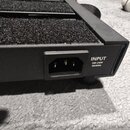 Detail zadní strany černého zařízení s texturou koberce na horní části. Je zde vidět napájecí konektor s nápisem INPUT 100-240V 50/60Hz. Povrch je v…