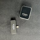 Dvě části bezdrátového systému značky SYNCO v černé barvě. Větší hranatý vysílač nese nápis SYNCO)) Transmitter a menší obdélníkový přijímač s konekt…