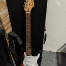 Elektrická kytara s tónem sunburst a bílým pickguardem. Na hlavě hředu je viditelné logo Fender a nápis STRATOCASTER. Kytara má hnědý hmatník a je op…