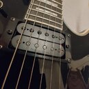 Detail elektrické kytary s černým lakovaným povrchem a hnědým hmatníkem. Na kytaru jsou instalovány dva humbuckery, z nichž na spodním je viditelný n…
