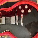 Černá elektrická kytara s tvarem singlecut v červeném plišovém kufru. Nástroj má dva humbuckery, čtyři ovládací knoflíky a černý plechtrguard. Na těl…