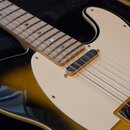 Detail elektrické kytary s tělem v provedení sunburst a bílým pickguardem. Na hrazence jsou vidět kovové pračky a modré markery v hřbitu. Kytara má j…