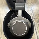 Šedé sluchátka s černými mušlami v černém přepravním kufru. Na horní části je bílý nápis beyerdynamic)))) a nad ním černá větřná clona. V pozadí je v…