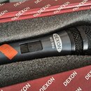 Černý dynamický mikrofon s mřížkou v horní části, umístěný v černé pěnové výplni červeného kufru. Na těle mikrofonu je bílý oválný štítek s nápisem D…