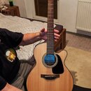 Akustická kytara s vyřezem v těle, přírodní dřevěnou barvou a černým plektrem. Na hlavě je viditelné logo Takamine. U otvoru rezonátoru je vložena mo…