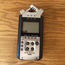 Stříbrno-černý digitální záznamník s nápisy ZOOM v horní části a Handy Recorder H4n ve spodní části. Zařízení má LCD displej, ovládací tlačítka a v h…
