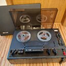 Černý cívkový magnetofon s průhledným plastovým krytem. Na přední straně je bílý nápis B 700 MADE IN CZECHOSLOVAKIA a červené logo v kruhu. Vlevo na…