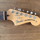 Fotografie zachycuje hlavu elektrické kytary z přírodního světlého dřeva s hnědým hmatníkem. Na hlavě je černý nápis Fender® JAZZMASTER®. Jsou zde vi…