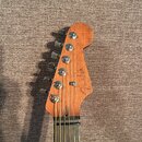 Fotografie zachycuje hlavu elektrické kytary z přírodního dřeva s šesti chromovanými ladičkami. Na povrchu je vidět nápis Fender v kurzívě. Hředen je…