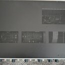 Horní strana šedého kovového zařízení s matným povrchem. V horní pravé části a v dolní polovině jsou umístěny ventilační mřížky s drobnými otvory. Na…