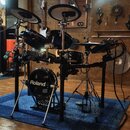 Elektronická bicí souprava značky Roland v černé barvě, umístěná na modrém koberci. Na basovém bubnu je bílý nápis Roland a níže označení V-Drums. So…