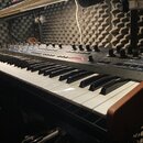 Černý syntetizátor s klaviaturaí a řadou ovládacích knoflíků na horním panelu. Nástroj má dřevěné boční panely v hnědé barvě. Na ovládacím panelu je…