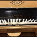 Světle hnědé dřevěné piano s černým povrchem nad klávesnicí. Uprostřed černého panelu je zlatý nápis A.PROKSCH. Klávesnice vykazuje známky opotřebení…