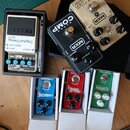 Fotografie zobrazuje šest kytarových efektů. V horní části jsou MXR Super COMP v černé barvě, MXR Custom Badass Modified OD v zlatovém provedení a BO…